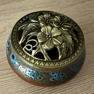 May include: A brown ceramic incense burner with a blue and brown glaze. The burner has a metal lid with a floral design.