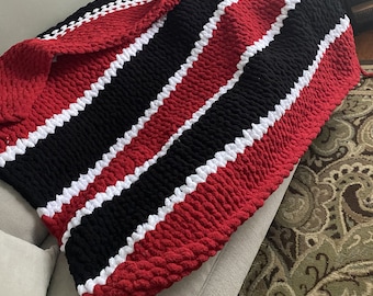 May include: A red, black, and white striped crocheted blanket with a textured, puffy design. The blanket is folded and draped over a couch.