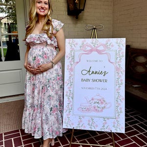 May include: A white sign with pink floral accents and a gold frame announces "Welcome to Annie's Baby Shower" with the date "November 17th, 2024". A teapot with a pink floral design is pictured on the sign. The sign is on a gold easel.