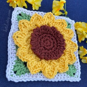 May include: Crocheted sunflower square with a brown centre, yellow petals, and green leaves. The square is surrounded by a white border.