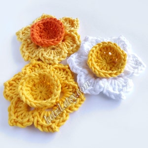 Op de afbeelding: Drie gehaakte narcissenbloemen. Eén bloem is wit, één is geel en één is geel met een oranje hart.