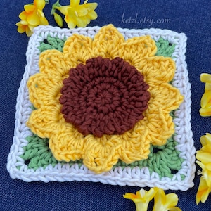 May include: A crocheted sunflower square with a brown center, yellow petals, and green leaves. The square is surrounded by a white border.