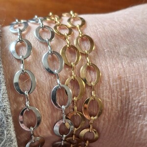 May include: Three bracelets with oval links, one in silver and two in gold, are displayed. The bracelets are worn on a wrist, showcasing their design and color variations. The links are connected in a chain-like pattern.