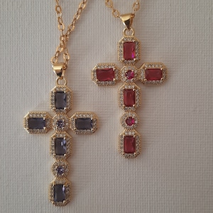 Peut inclure: Deux pendentifs en forme de croix dorées ornés de pierres précieuses rectangulaires et rondes. L'une des croix présente des pierres rouges, l'autre des pierres violettes. Les deux croix sont sur de fines chaînes dorées, sur fond blanc.