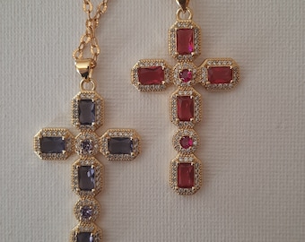 Puede incluir: Dos colgantes de cruz dorados con piedras preciosas rectangulares y redondas. Una cruz presenta piedras rojas, mientras que la otra tiene piedras moradas. Ambas cruces están sobre delicadas cadenas doradas, sobre un fondo blanco.