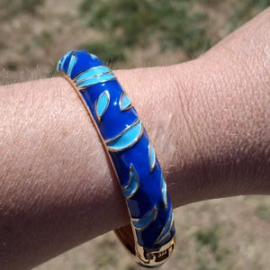 May include: A blue enamel bangle bracelet with a gold-tone base. The bracelet features a repeating pattern of blue and light blue leaf-like shapes. The bracelet is on a wrist.