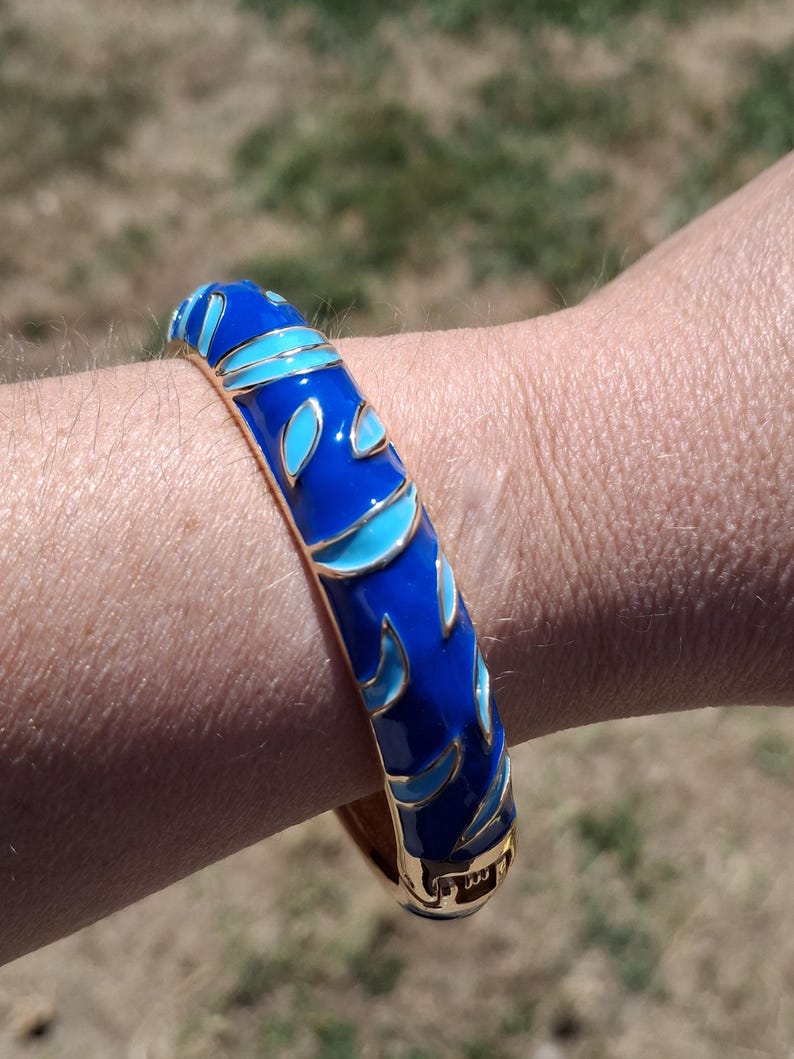 May include: A blue enamel bangle bracelet with a gold-tone base. The bracelet features a repeating pattern of blue and light blue leaf-like shapes. The bracelet is on a wrist.