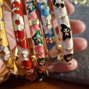 May include: A collection of colorful enamel bangles. The bangles feature floral designs in red, white, blue, pink, and black, with gold accents. The bangles are a mix of colors and patterns.