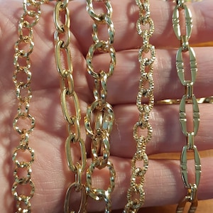 Puede incluir: Cinco cadenas de oro de diferentes estilos y grosores. Las cadenas están dispuestas en fila sobre una mano.