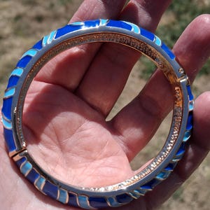 May include: A hinged bangle bracelet with a gold-tone interior and a silver-tone exterior. The bracelet features a repeating pattern of blue enamel in various shades. The bracelet is approximately 2.5 inches in diameter.