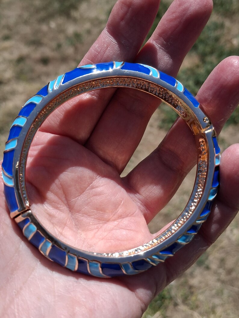 May include: A hinged bangle bracelet with a gold-tone interior and a silver-tone exterior. The bracelet features a repeating pattern of blue enamel in various shades. The bracelet is approximately 2.5 inches in diameter.