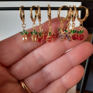 Puede incluir: Un conjunto de seis pendientes de aro de oro con colgantes. Cada colgante presenta una fruta diferente: una cereza, una piña, una uva y una fresa. Los colgantes están hechos de oro y tienen piedras preciosas de colores.