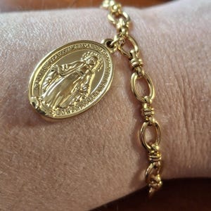 Puede incluir: Pulsera dorada con un colgante ovalado que representa una figura religiosa y una inscripción. La pulsera tiene un diseño de cadena delicado con un cierre de mosquetón. El colgante y la cadena son de color dorado brillante.