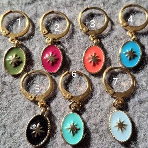 Puede incluir: Un conjunto de siete pendientes de aro dorados. Cada pendiente presenta un pequeño colgante ovalado con un diseño de estrella en varios colores: verde, rosa, naranja, azul, negro y azul claro. Los pendientes se muestran sobre una superficie gris.