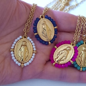 Peut inclure: Quatre colliers dorés avec des pendentifs ovales représentant la Médaille Miraculeuse. Chaque pendentif est entouré de petites perles blanches, bleu foncé, rose vif ou turquoise. Les colliers sont fixés à de fines chaînes en métal doré.