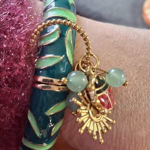 Puede incluir: Una pulsera de esmalte turquesa con detalles dorados y un dije. El dije presenta un sol dorado, una mariquita con detalles rojos y negros y cuentas verde claro. La pulsera tiene un diseño de hojas.