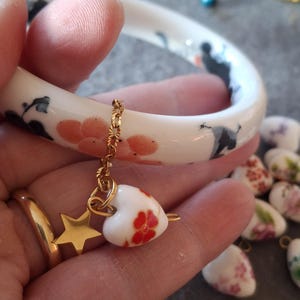 Peut inclure: Un bracelet jonc en céramique blanche avec un motif floral rose et noir. Une chaîne dorée suspend un charm en forme de cœur avec des fleurs rouges et une étoile dorée. D'autres breloques en forme de cœur sont visibles.