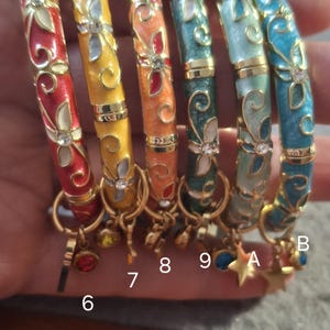 May include: Six hinged bangle bracelets in red, yellow, orange, dark green, light green, and blue, each featuring gold-tone floral filigree, embedded clear crystals, and a small dangling charm. The bracelets are labelled with numbers 6, 7, 8, 9 and letters A, B.