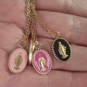 Puede incluir: Collares dorados con colgantes ovalados que representan una figura religiosa. Los colgantes tienen fondos rosas y negros con detalles dorados. Los collares se muestran sobre un fondo neutro.