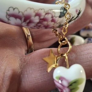 Könnte beinhalten: Weißes Keramikarmband mit floralem Design in Rosa und goldenen Akzenten. Eine goldene Kette mit einem Stern- und Herzanhänger hängt am Armband. Der Herzanhänger zeigt ein rosa Blumenmuster.