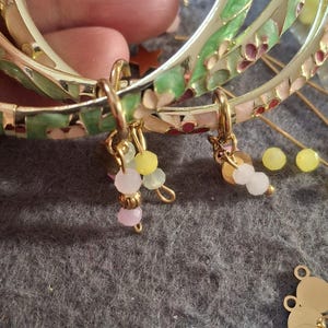 Könnte beinhalten: Goldfarbene Armreifen mit floralen und Blattmustern in Grün, Rosa und Rot. Jeder Armreif hat kleine, baumelnde Anhänger mit bunten Perlen in Rosa, Gelb und Weiß. Der Schmuck ist vor einem grauen Hintergrund platziert.