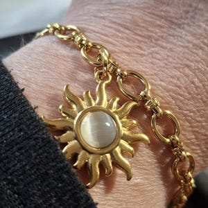 Peut inclure: Bracelet doré orné d'un charm en forme de soleil avec une pierre centrale ovale blanc cassé. Le motif soleil est détaillé, et le bracelet est composé d'une chaîne à maillons. Le bracelet est porté sur un poignet, avec une manche noire visible.
