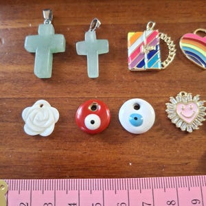 Peut inclure: Une collection de breloques et de pendentifs de bijoux. Comprend deux croix en pierre vert clair, un charm livre en émail coloré, un cœur arc-en-ciel, une rose blanche, des perles rouges et blanches en œil de mauvais œil et un soleil doré souriant. Un mètre ruban est visible.