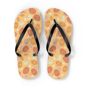 May include: A pair of flip-flops with a pumpkin and leaf pattern on a light orange background. The flip-flops have black straps and a fall theme. The design includes orange pumpkins and various leaves.