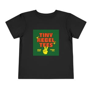 May include: Black toddler t-shirt with a green square graphic. The graphic reads "TINY REBEL TEES" in red letters, with a yellow peace sign and the phrases "SPEAK GOOD" and "FORBID EVIL".