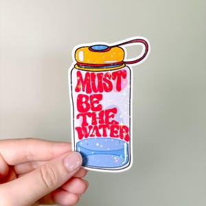 Puede incluir: Una pegatina de botella de agua blanca con una tapa amarilla y un texto rojo y blanco que dice "Must Be The Water".