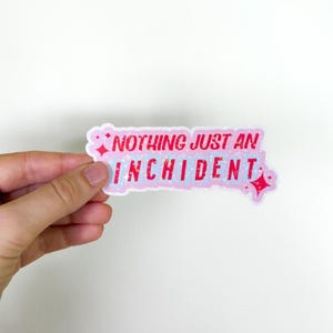 以下が含まれることがあります： ピンクと白のキラキラしたステッカーに、赤い文字で「Nothing just an incident」と書かれています。