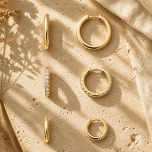 May include: A selection of gold hoop earrings, some plain and others adorned with diamonds. The earrings are arranged on a textured, light-toned surface, with shadows adding depth. The image presents a variety of gold hoop styles and sizes, suitable for different tastes.