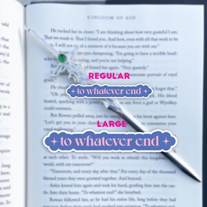 May include: A close-up of a book with the text "Kingdom of Ash" on the cover. A silver bookmark with a green gem is placed on the page. The bookmark has two blue and pink stickers with the text "to whatever end" on them. The text on the stickers is in a bold, white font.
