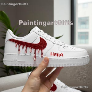 Può includere: Sneaker bianca con uno swoosh Nike rosso tempestato di gioielli e un design a goccia. La parola "Hana" è scritta in rosso sul lato. La scarpa è tenuta in mano.