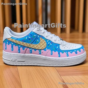 May include: White and blue sneaker with a unique ice cream cone design. The shoe features a pink base with blue dripping accents, a waffle cone-shaped logo, and colorful sprinkles. The word "AIR" is visible on the side.