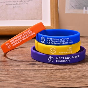 Può includere: Un assortimento di braccialetti di allerta medica in vari colori. I braccialetti includono uno arancione con testo che indica malattie cardiache e uno blu, giallo e viola con informazioni mediche e dettagli di contatto di emergenza. I braccialetti sono in silicone.