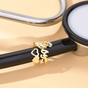 Peut inclure: Breloque de stéthoscope en or avec le mot "Infirmière" et un cœur. Le breloque a une petite pierre précieuse bleue.