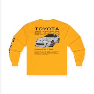 May include: Mustard yellow long-sleeve shirt featuring a graphic of a silver Toyota Supra MK IV car. The text "TOYOTA SUPRA" is above the car graphic, with additional text below. The sleeve has a black graphic.