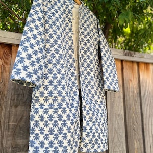 May include: A long, white quilted jacket with a blue floral pattern. The jacket has a button closure and long sleeves.