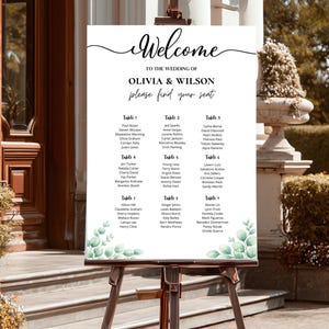 Puede incluir: Un letrero de bienvenida de boda blanco sobre un caballete de madera. El letrero dice "Bienvenida" encima de los nombres "Olivia & Wilson" y un plano de asientos con los nombres de los invitados. Ilustraciones de hojas verdes adornan las esquinas inferiores.