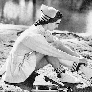 May include: A woman wearing a white sweater and a striped knit hat sits on a snowy surface with her ice skates on. She is wearing white socks with black stripes.