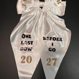 May include: A white satin hair bow featuring gold glitter text that reads "SENIOR" on the top loops. The long tails display black text reading "ONE LAST BOW" and "BEFORE I GO" with the gold glitter numbers "20" and "27" at the bottom.