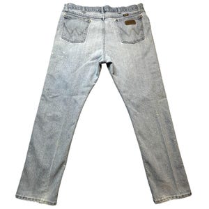 May include: Light blue denim jeans with a classic five-pocket design. The jeans feature a brown leather patch and the Wrangler logo on the back pocket. The jeans are pre-worn and have some minor wear and tear.