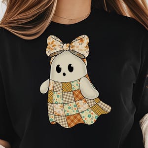 Pode incluir: Camiseta preta com um desenho de fantasma de desenho animado. O fantasma tem um corpo creme, olhos pretos e um la&ccedil;o com padr&otilde;es de outono. O design de patchwork inclui padr&otilde;es florais e xadrez em tons de laranja, marrom e amarelo.