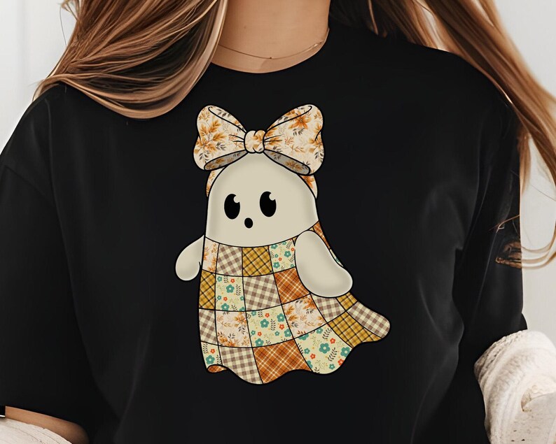 Pode incluir: Camiseta preta com um desenho de fantasma de desenho animado. O fantasma tem um corpo creme, olhos pretos e um la&ccedil;o com padr&otilde;es de outono. O design de patchwork inclui padr&otilde;es florais e xadrez em tons de laranja, marrom e amarelo.