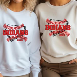 May include: Two white sweatshirts with a baseball-themed graphic. The graphic features a red baseball diamond with the word "INDIANS" in red text and the year "2023" in black text. The diamond is surrounded by red and black baseball stitching.