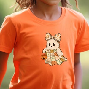 Pode incluir: Camiseta laranja com um desenho de fantasma de desenho animado. O fantasma usa um la&ccedil;o, um vestido de retalhos e uma cesta de ab&oacute;bora. O design usa cores bege, laranja e amarelo, perfeito para o Halloween.