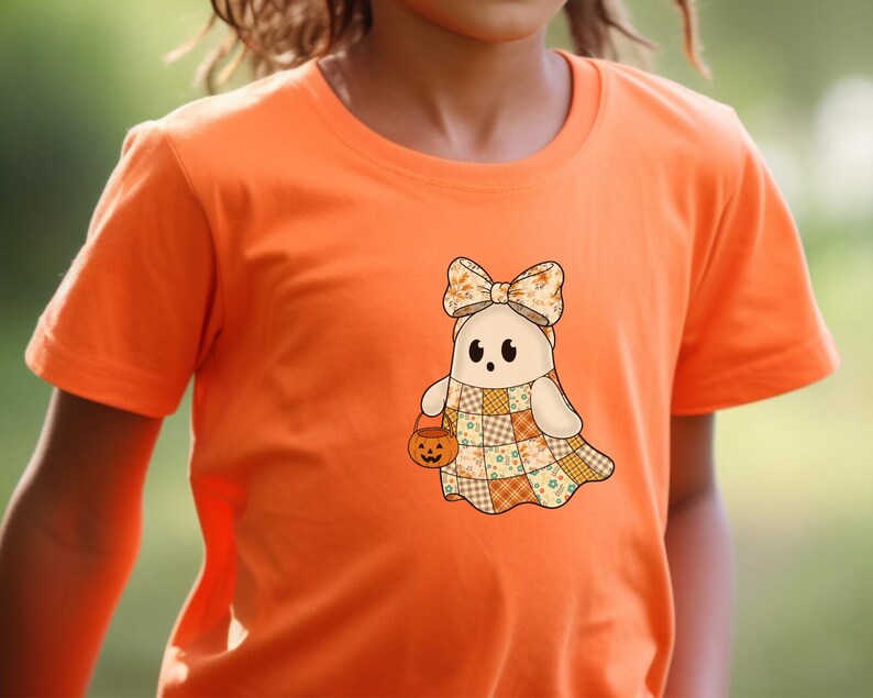 Pode incluir: Camiseta laranja com um desenho de fantasma de desenho animado. O fantasma usa um la&ccedil;o, um vestido de retalhos e uma cesta de ab&oacute;bora. O design usa cores bege, laranja e amarelo, perfeito para o Halloween.