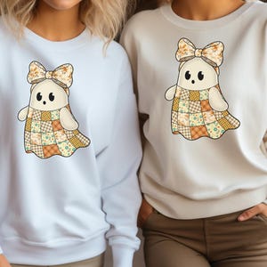 Pode incluir: Duas camisolas brancas e creme com um desenho de fantasma. O fantasma tem um vestido de retalhos, um la&ccedil;o floral e olhos pretos. As camisolas s&atilde;o de manga comprida, ideais para o outono ou Halloween.