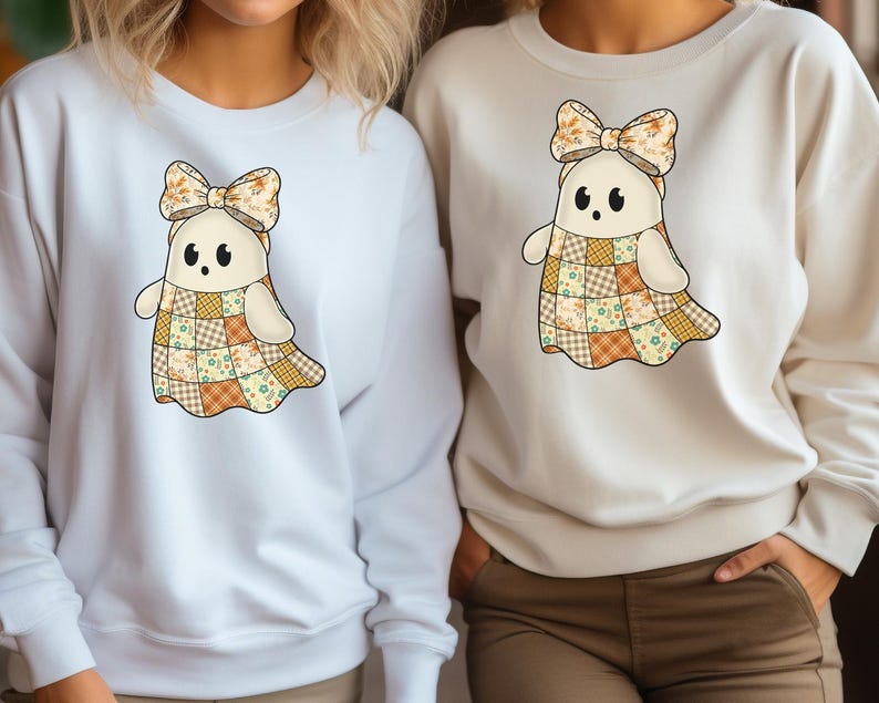 Pode incluir: Duas camisolas brancas e creme com um desenho de fantasma. O fantasma tem um vestido de retalhos, um la&ccedil;o floral e olhos pretos. As camisolas s&atilde;o de manga comprida, ideais para o outono ou Halloween.